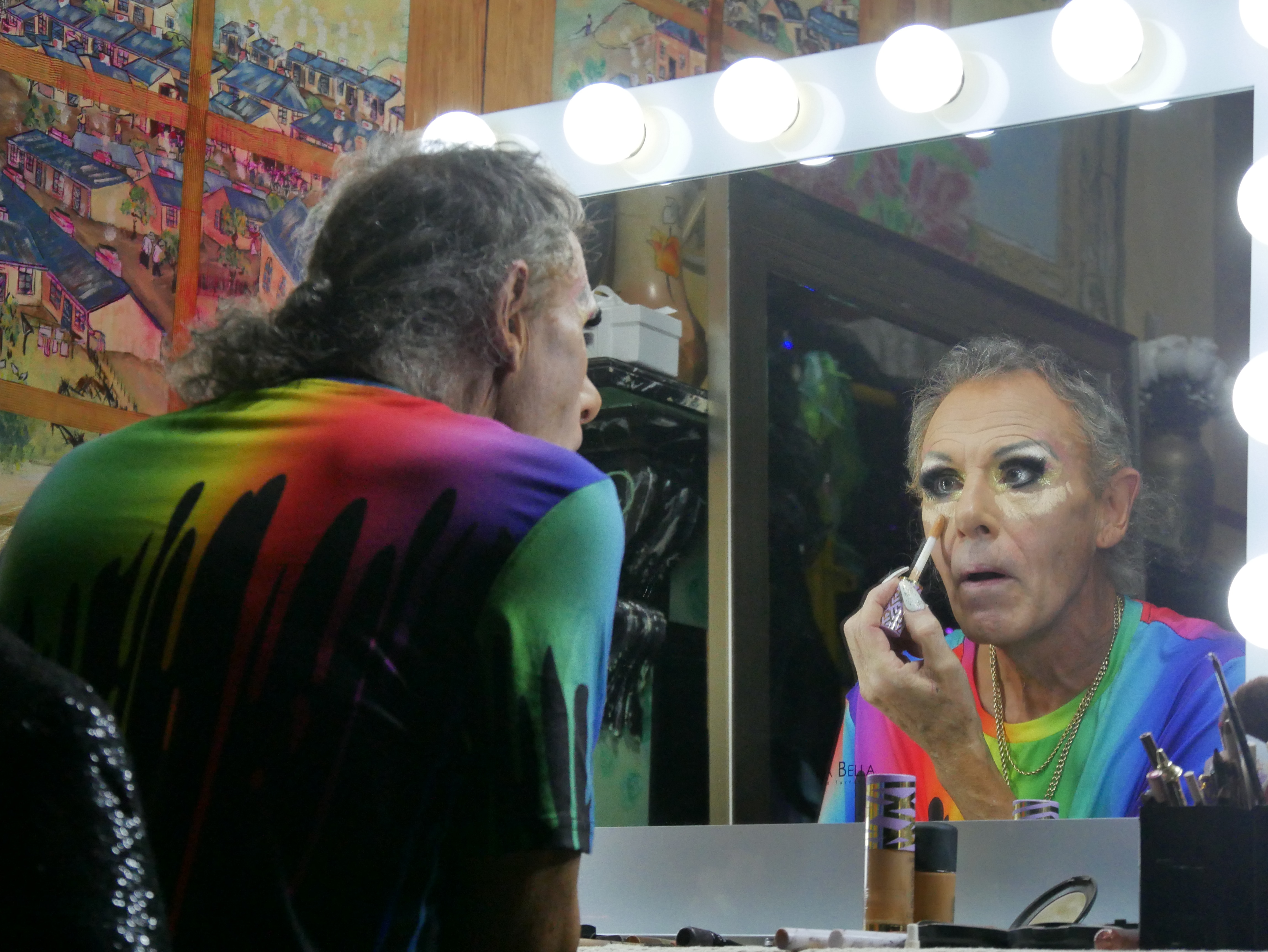 A drag queen wearing a rainbow tshirt sits and applied concealer in the mirror. 