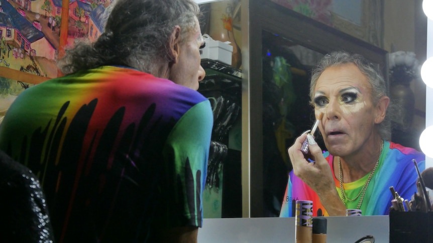 A drag queen wearing a rainbow tshirt sits and applied concealer in the mirror. 