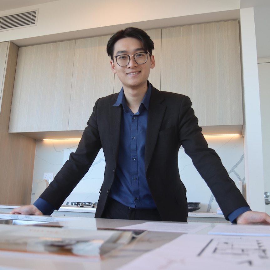 a man with short black hair and glasses wears a black suit jacket and leans on a new kitchen island table