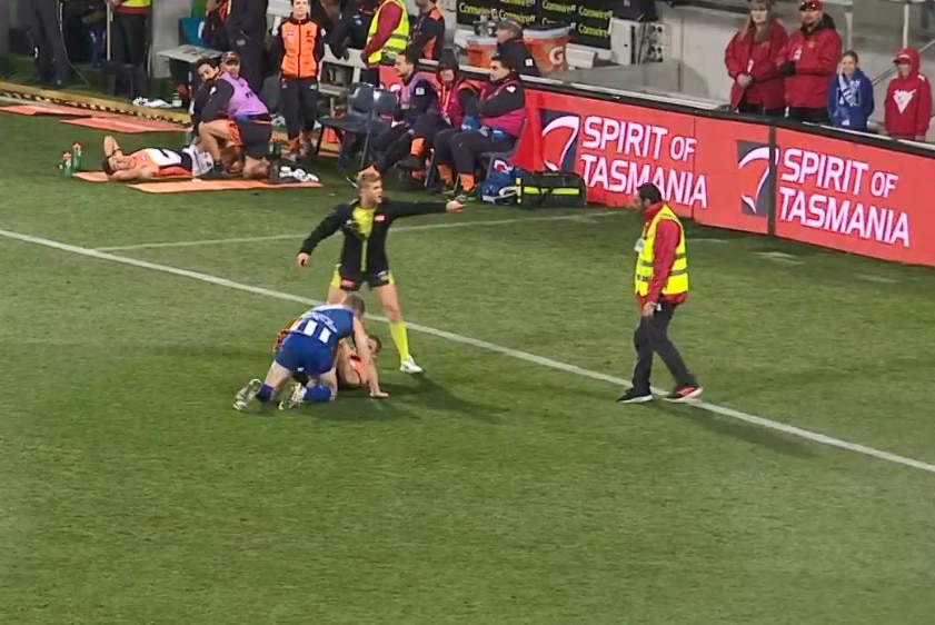 An umpire standing over two fighting AFL players points towards an approaching security guard.