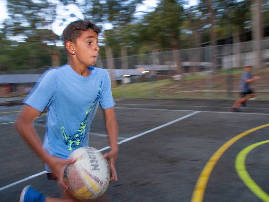 A boy in a light blue shirt runs past the camera holding a ball.