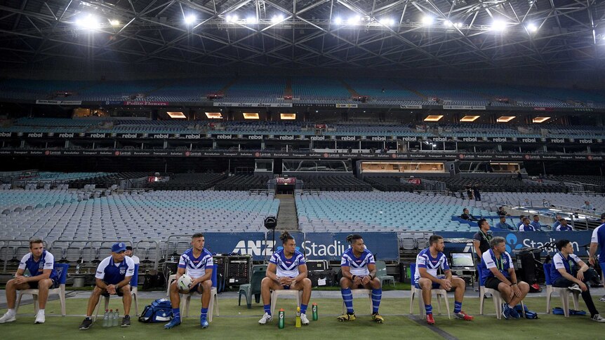 The Bulldogs NRL team's interchange players sit on a bench in front of an empty grandstand.