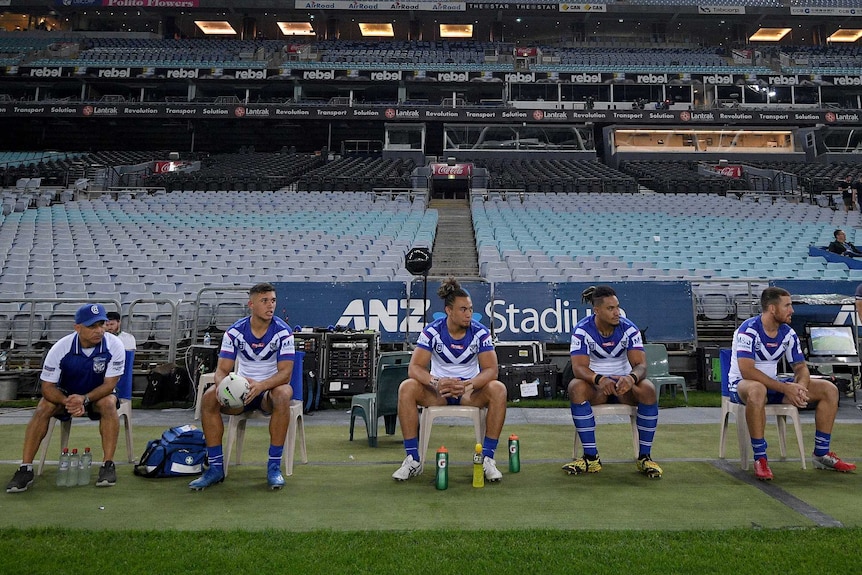 The Bulldogs NRL team's interchange players sit on a bench in front of an empty grandstand.