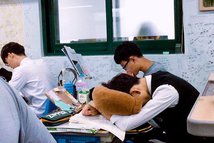 A young man with his head on a pillow at a desk while students study around him