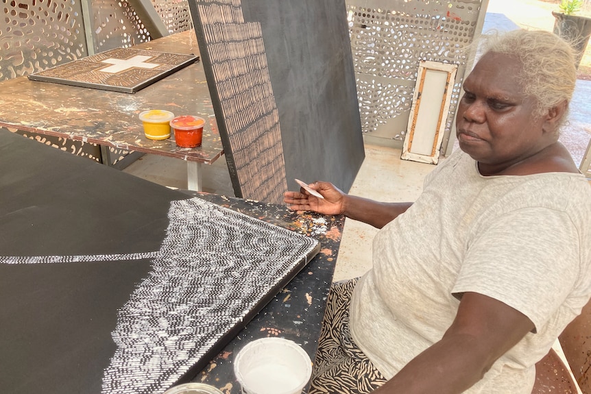 An blonde Aboriginal woman sits working with white paint on a corner of a large, dark canvas