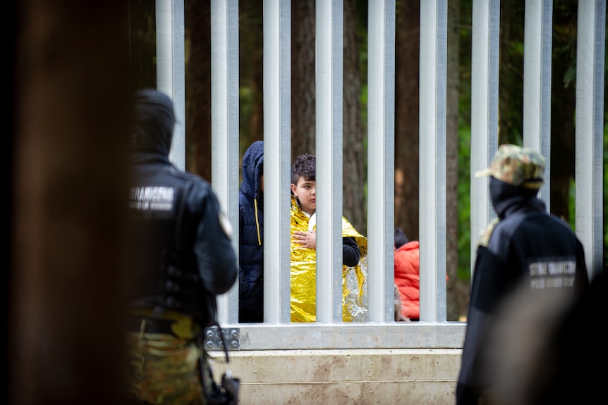 A young boy is seen through the fence wrapped in an emergency foil thermal blanket.  