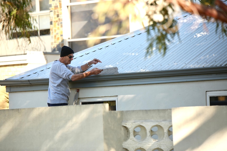 A man wearing a beanie and sunglasses installs gutter guard behind a wall.