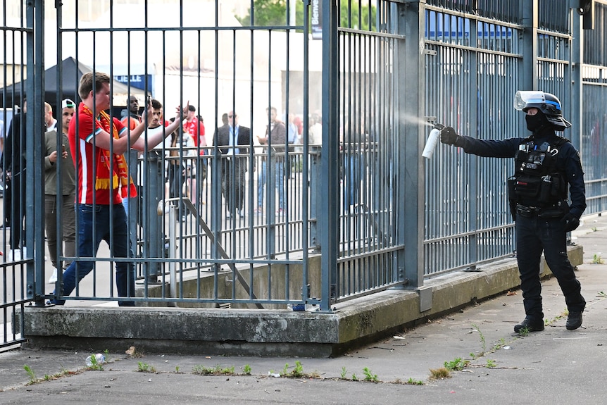 A riot police officer wearing a helmet and face covering uses an aerosol spray to send tear gas at a football fan.