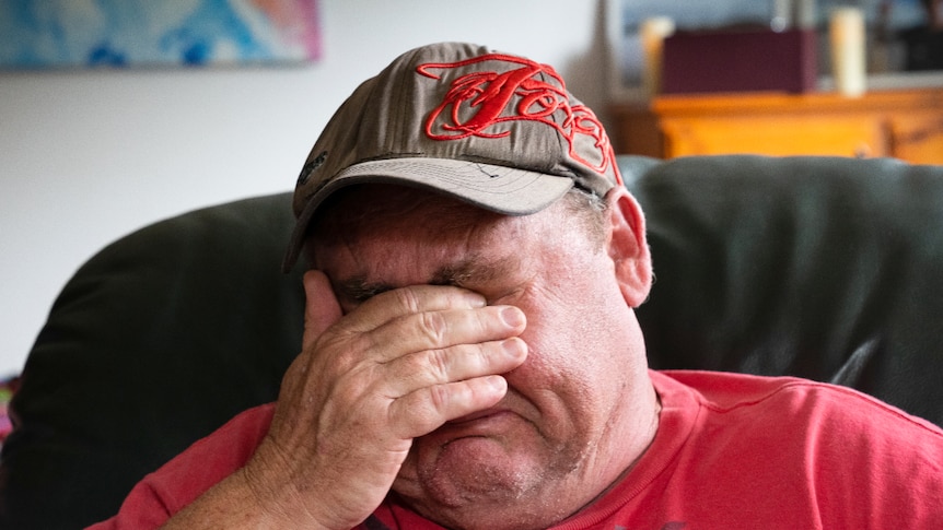A middle-aged man covers his face as he sits in a room, weeping.