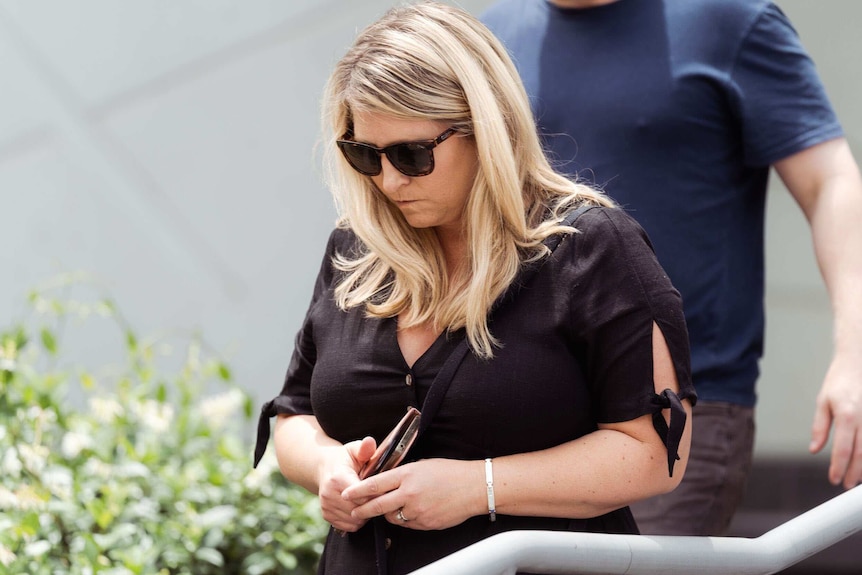 A blonde woman in dark sunglasses clutching a phone walks next to a railings.