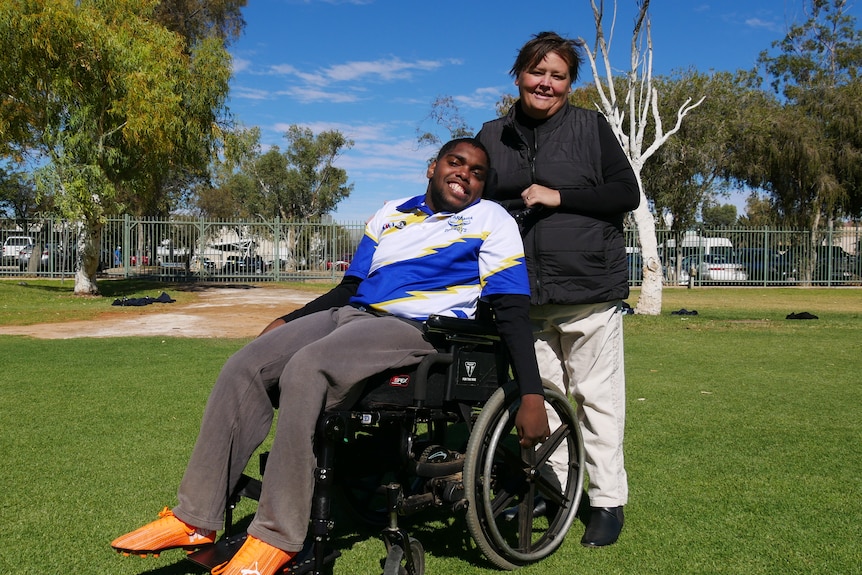 A man in a wheelchair with a woman standing behind him on a sports oval, both smiling.