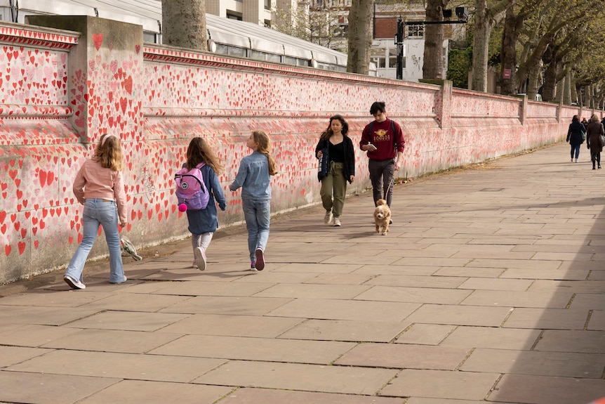 A group of three children play next to a wall decorated with red hearts as two women walk by.