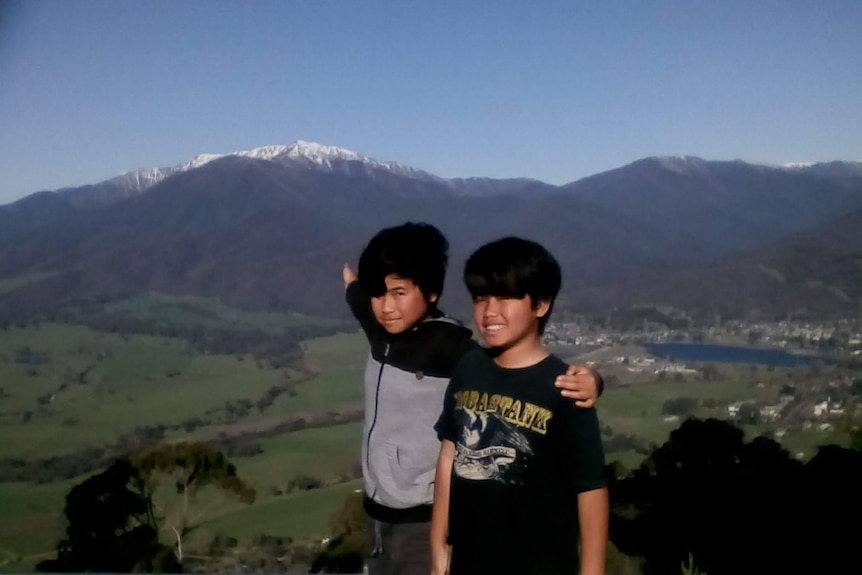 Two boys smiling at the camera against a backdrop of mountain ranges, one with snow.