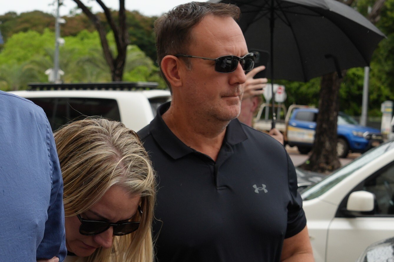 A white man, black polo shirt, sunglasses walking, blonde-haired woman crouched next to him