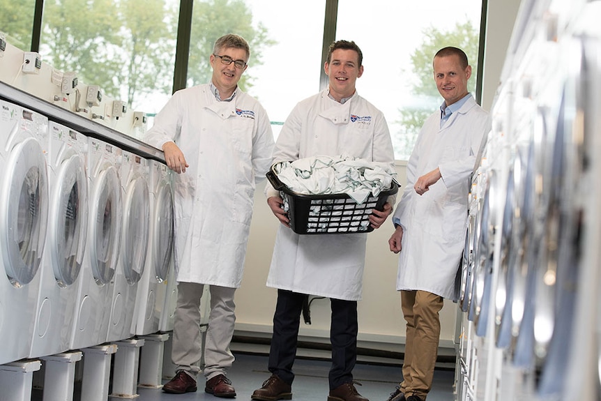 Grant Burgess, Max Kelly and Neil Lant stand near several washing machines holding washing baskets.