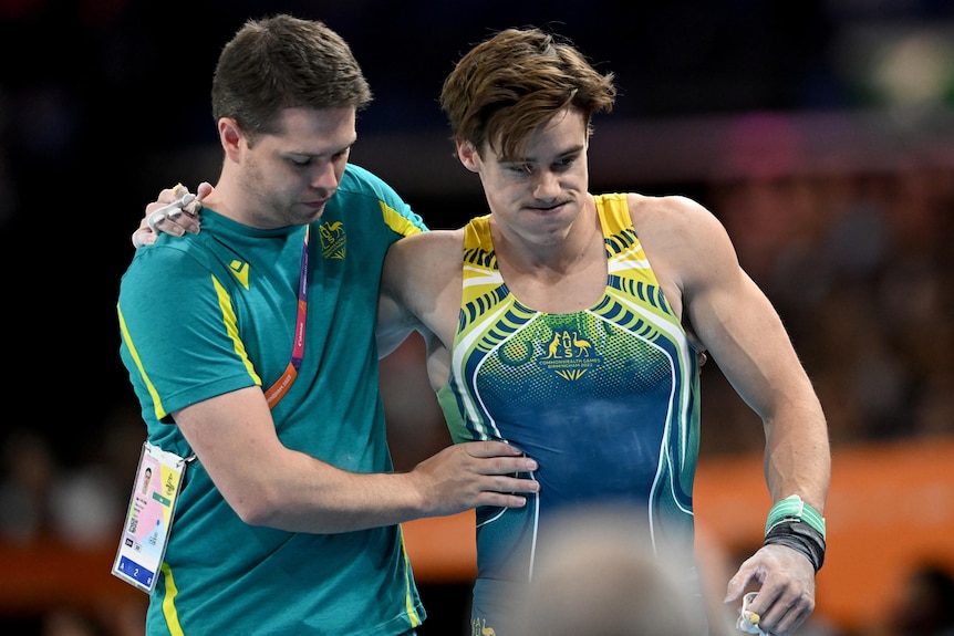 A trainer puts his arm around an Australian gymnast, who grimaces as he hobbles away after his horizontal bar routine.