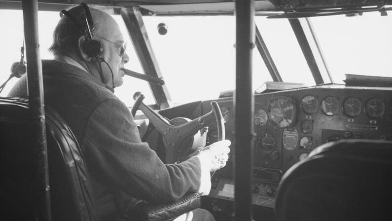 A large man wearing headphones and with a cigar in his mouth at the controls of an plane