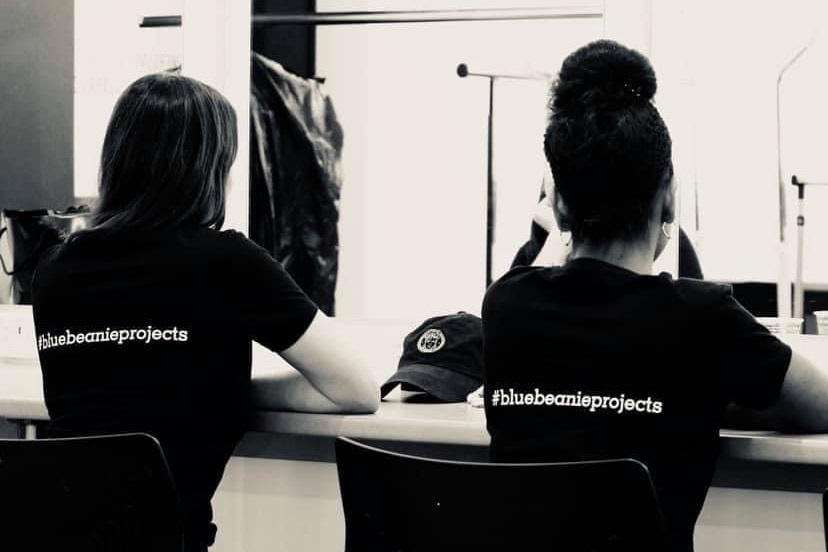 Two people sit at a table facing away from the camera, the backs of their T-shirts read #bluebeanieprojects