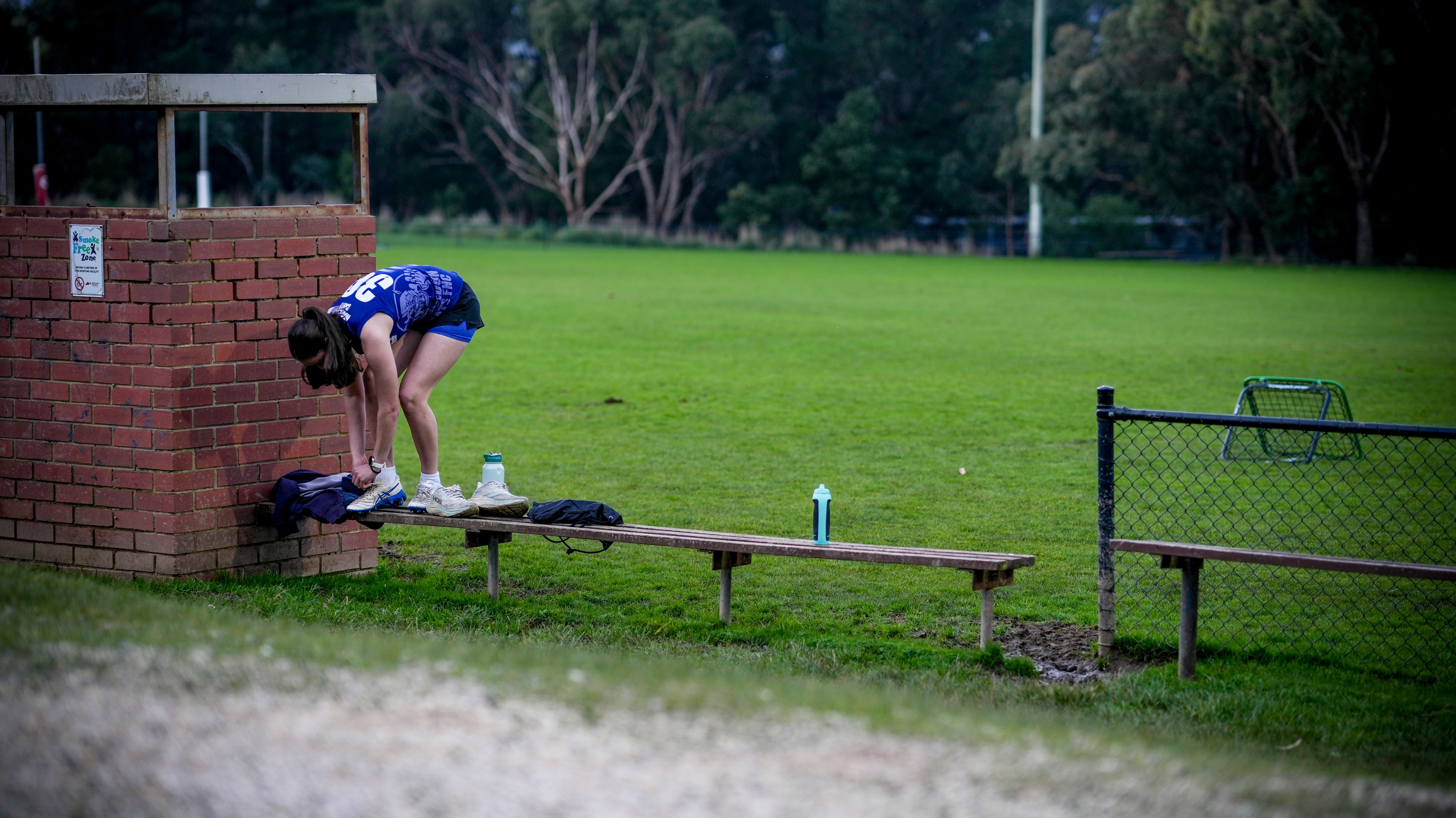 A woman in football gear stands on a bench to put on her shoes.
