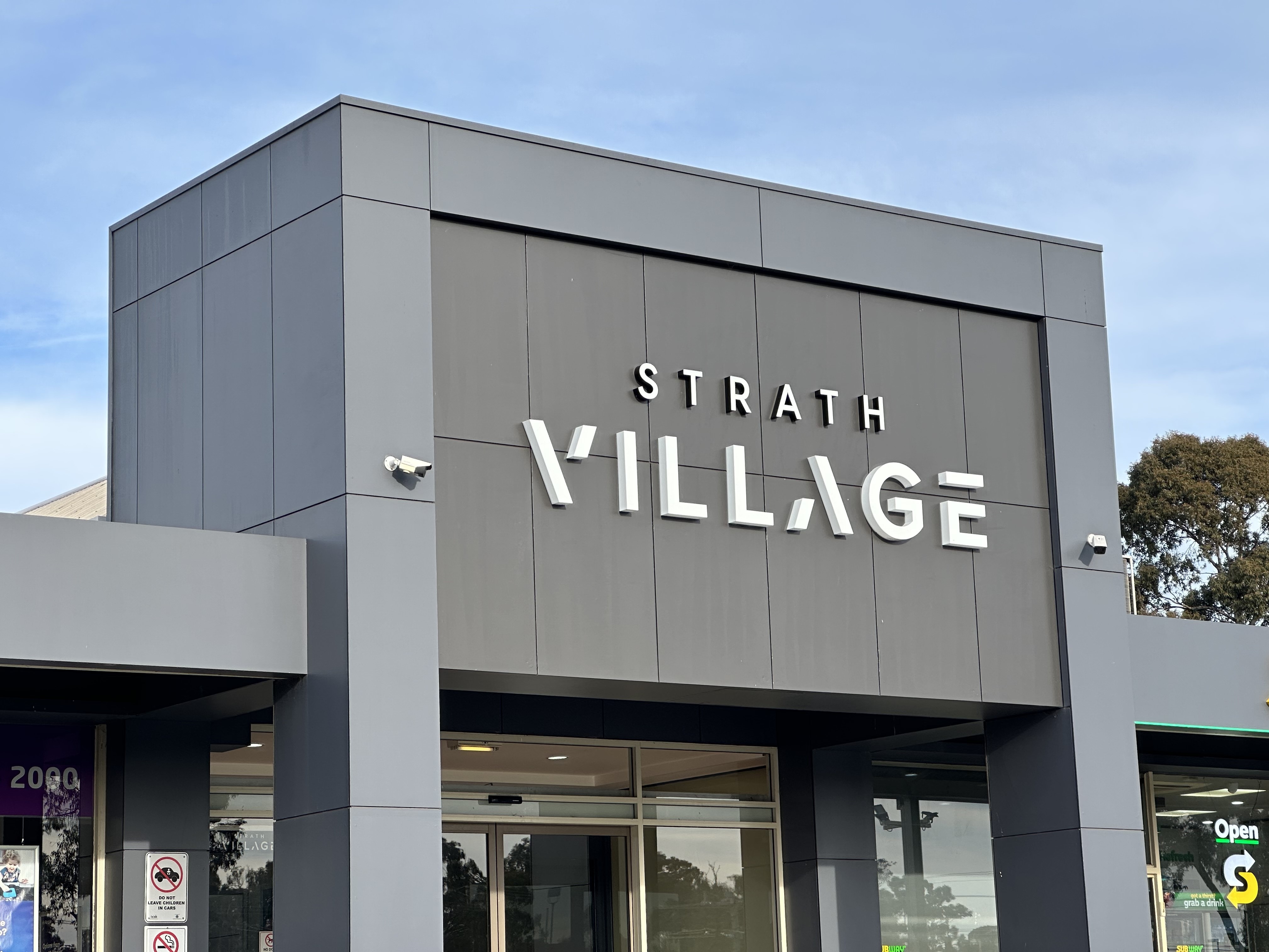 A square grey building with Strath Village sign, glass doors, light blue sky.