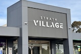 A square grey building with Strath Village sign, glass doors, light blue sky.
