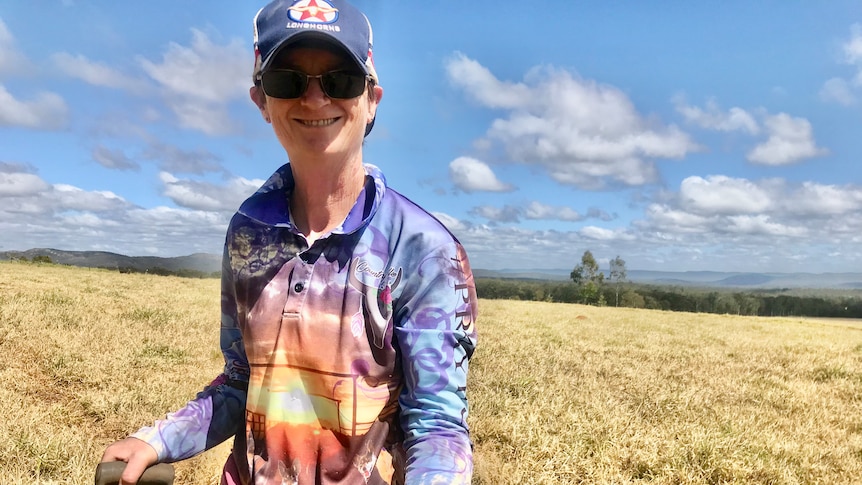 A woman with a hat and sunglasses stands in a paddock holding a shovel in one hand and a dung beetle in the other.