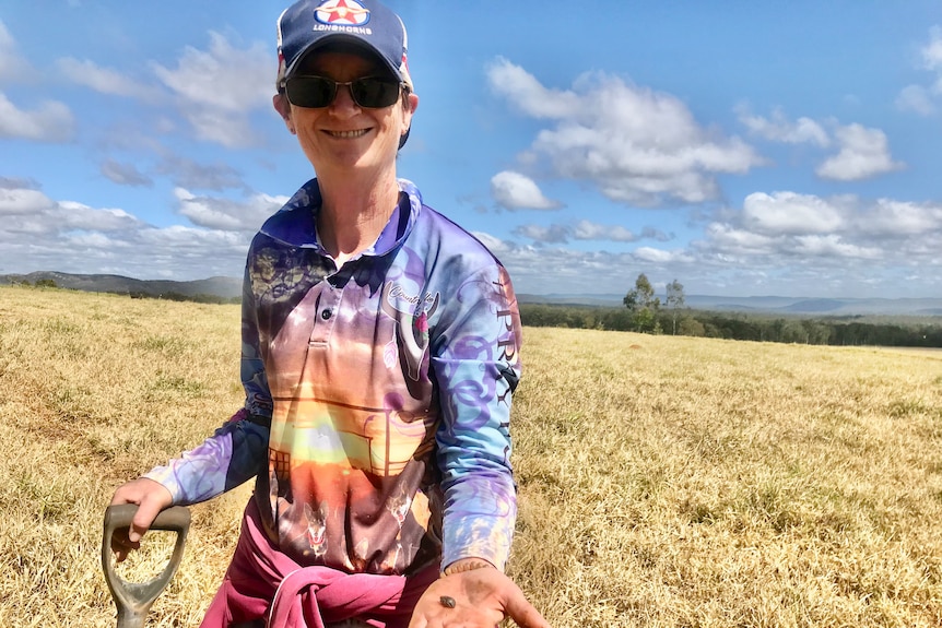 A woman with a hat and sunglasses stands in a paddock holding a shovel in one hand and a dung beetle in the other.