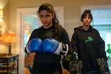 Young Pakistani woman with wavy black hair in a ponytail wears black gym clothes and blue boxing gloves, while a friend looks on