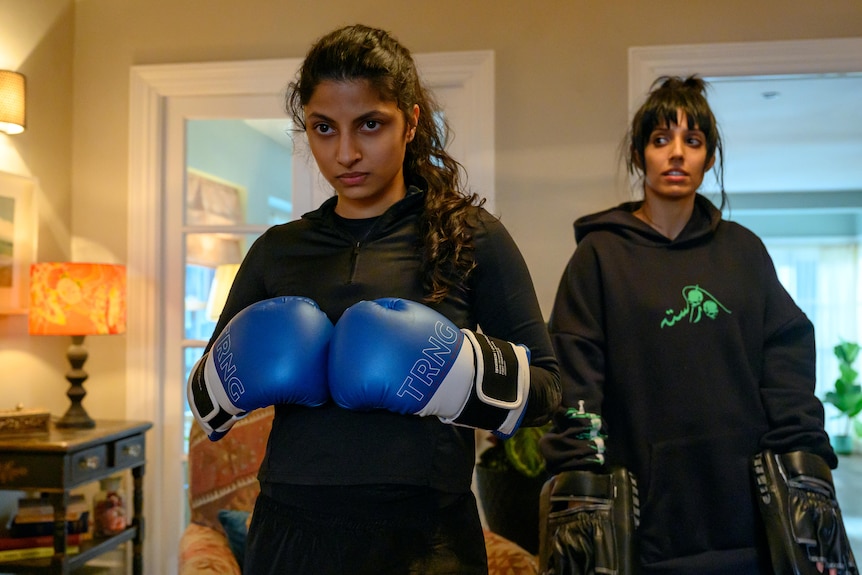 Young Pakistani woman with wavy black hair in a ponytail wears black gym clothes and blue boxing gloves, while a friend looks on