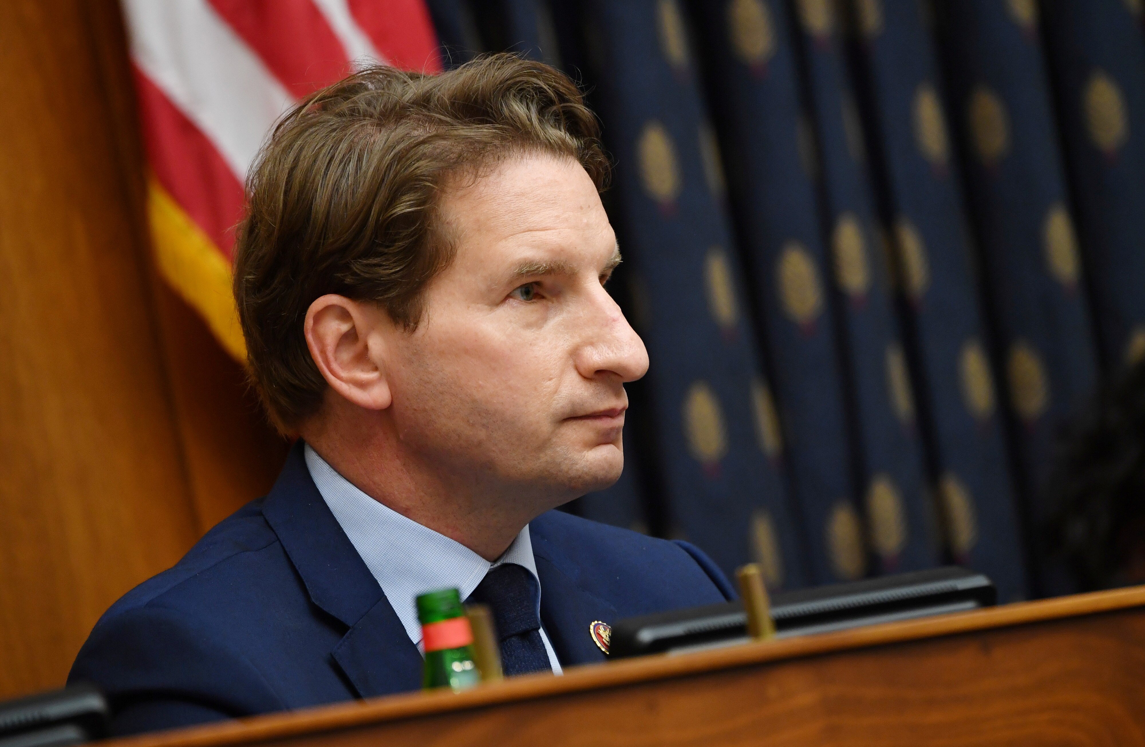 A middle-aged white man in a suit with light brown hair sits at a wood-panelled bench in front of an American flag.