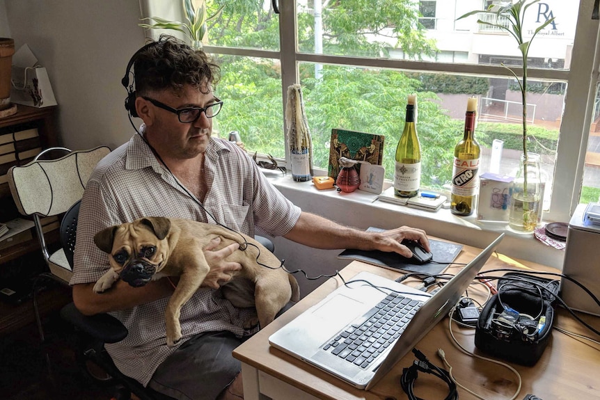 A middle-aged white man sits at a desk working on his computer, a dog in his lap
