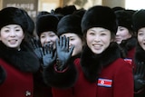 Young North Korean women wearing matching black hats and fur-trimmed red coats wave at the camera.