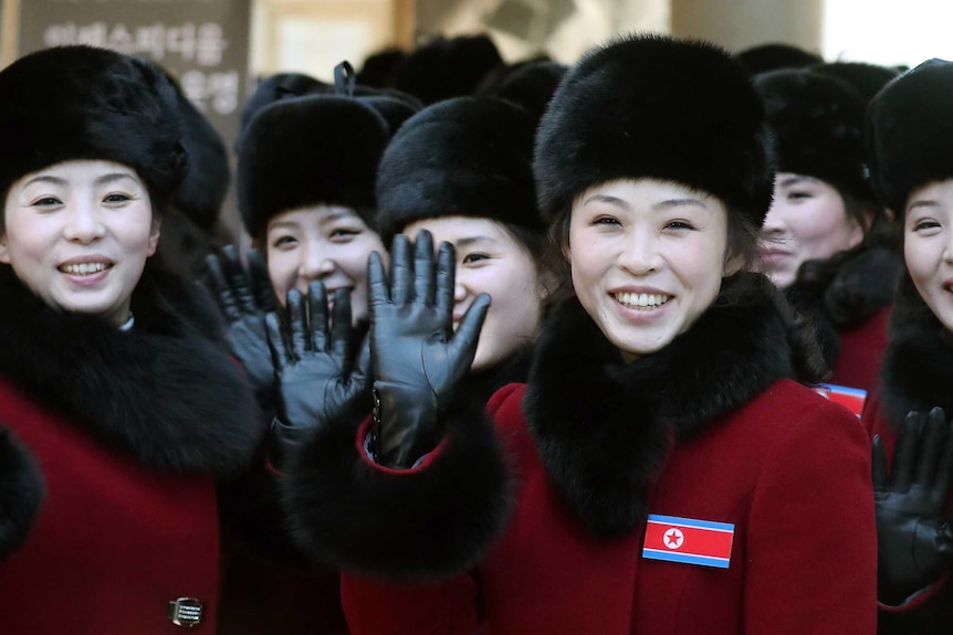 Young North Korean women wearing matching black hats and fur-trimmed red coats wave at the camera.