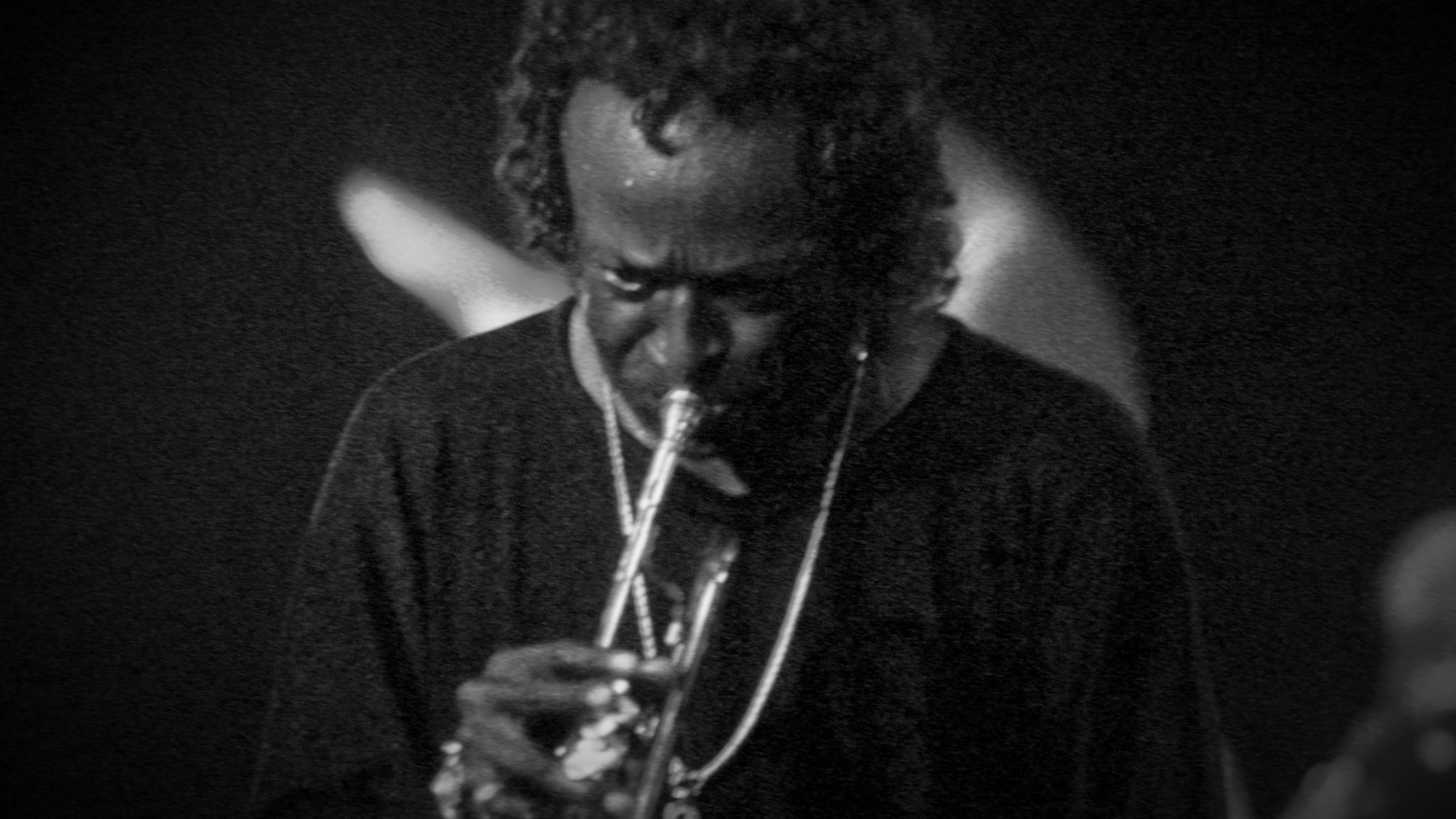 A monochrome, grainy photo of Miles Davis in the 1970s with long hair