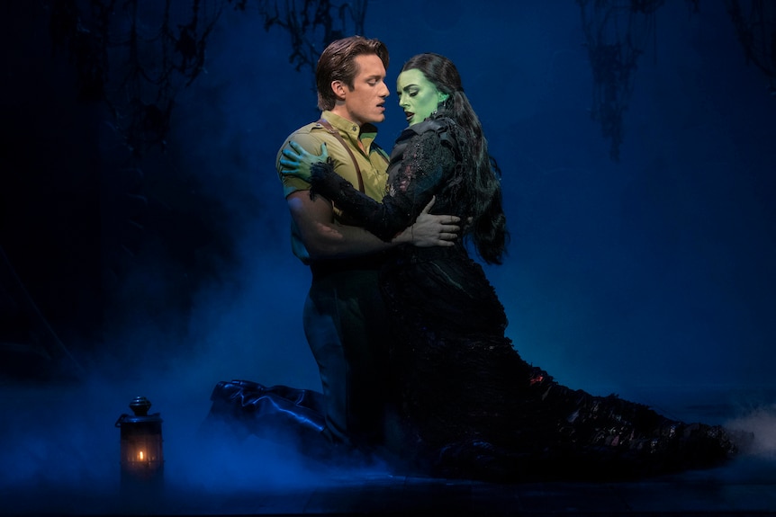 On stage, a young man grasps a woman with a painted green face and hands tightly, passionately, around the waist