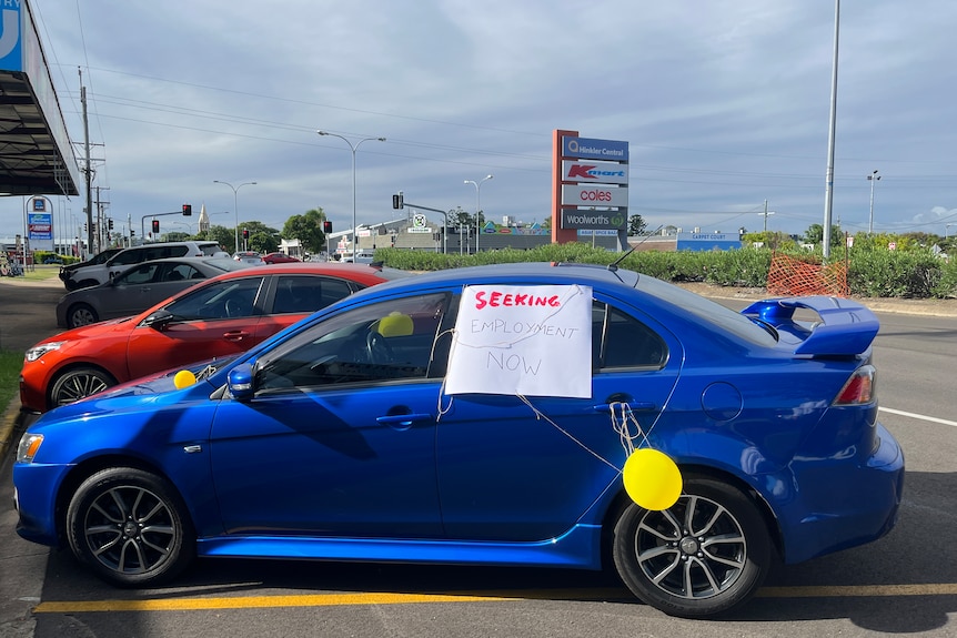 A blue sedan with a  yellow balloon on the door handle and a white sign which says 'Seeking employment now' in red writing. 