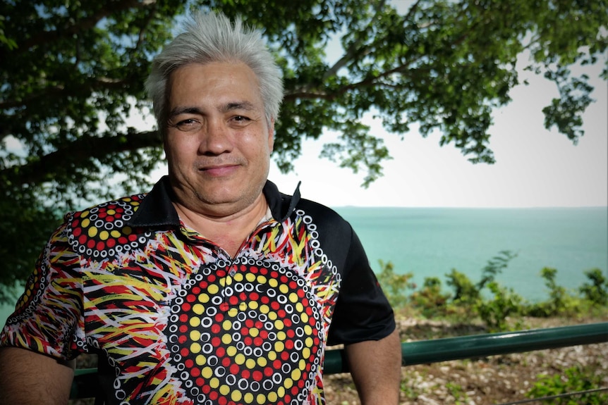 Mid-shot of man wearing Aboriginal print shirt standing on top of a cliff. Darwin Harbour in background