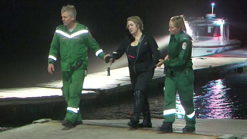 A woman wearing a black wetsuit has two paramedics next to her and a boat and water behind