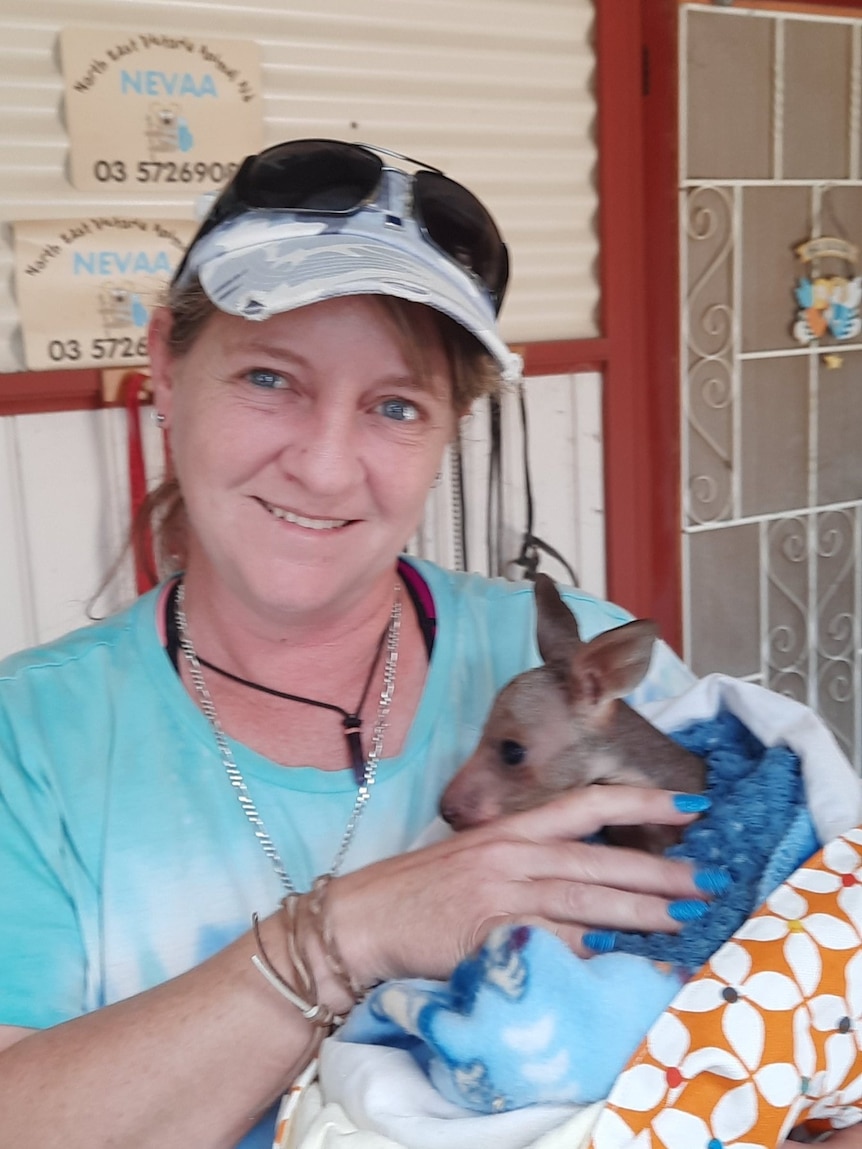 A woman wearing a cap smiles at the camera while holding a joey wrapped in a blanket