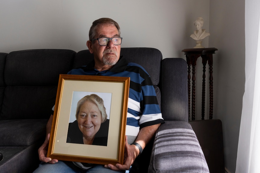 A man sits on a couch, holding a large framed portrait of his wife Gayle. In the photo Gayle is smiling.
