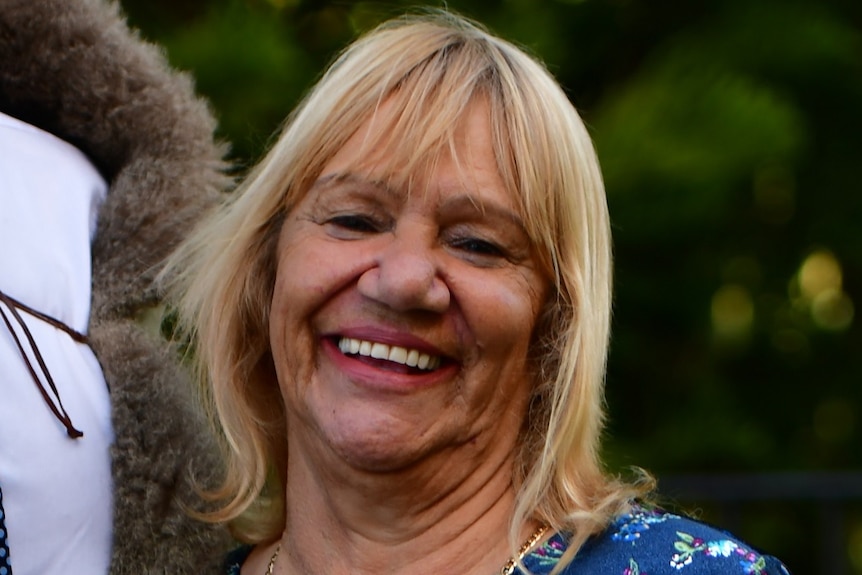 Aunty Geraldine Atkinson smiles as she stands outside, wearing a blue dress.