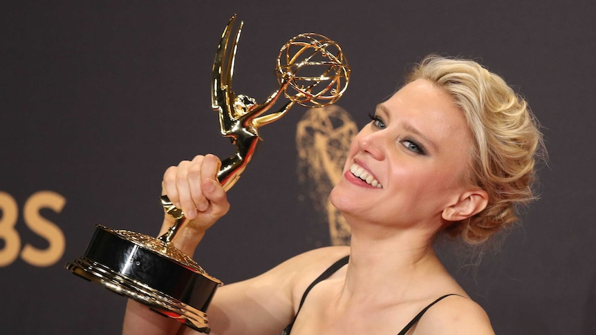 Kate McKinnon shows a wide toothy grin and holds her emmy high beside her face.