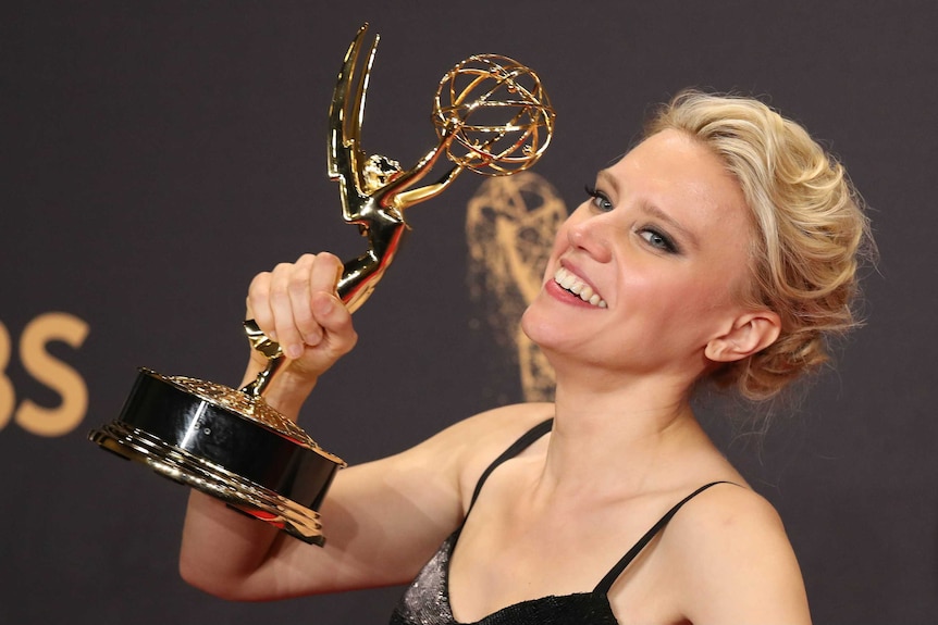 Kate McKinnon shows a wide toothy grin and holds her emmy high beside her face.
