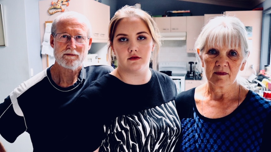 Young woman Amy Nivison-Smith stands between her parents in a kitchen.