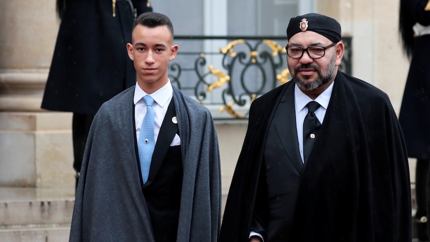 A man with a beard stand next to a younger man in a suit 