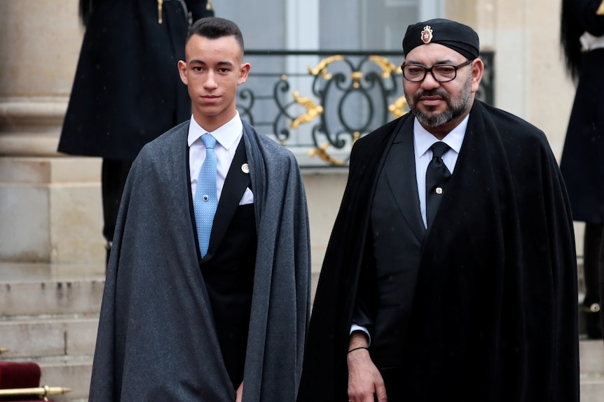 A man with a beard stand next to a younger man in a suit 