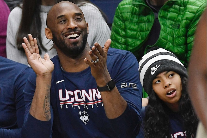 Kobe Byrant claps in a blue shirt while sitting in the crowd with his daughter Gianna wearing a beanie