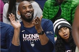Kobe Byrant claps in a blue shirt while sitting in the crowd with his daughter Gianna wearing a beanie