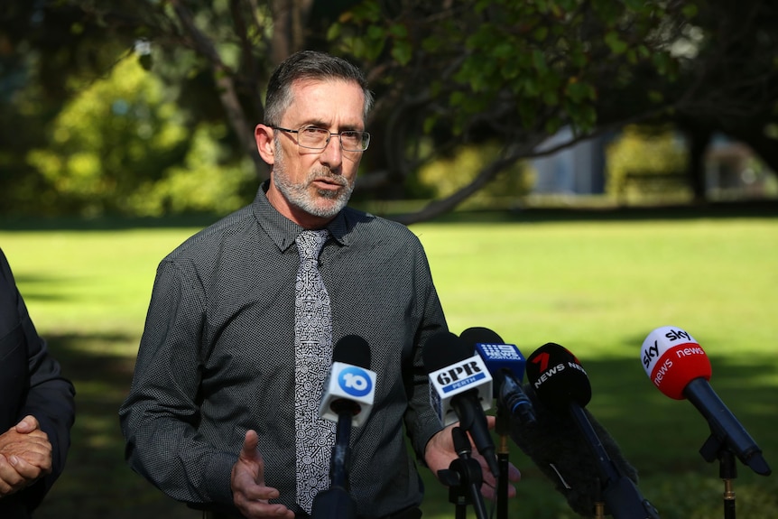 Dan wears a dark shirt and tie as he addresses media in a park setting