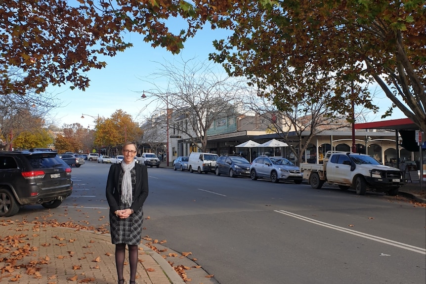 A woman stands by the side of the road in the main street of a country town.